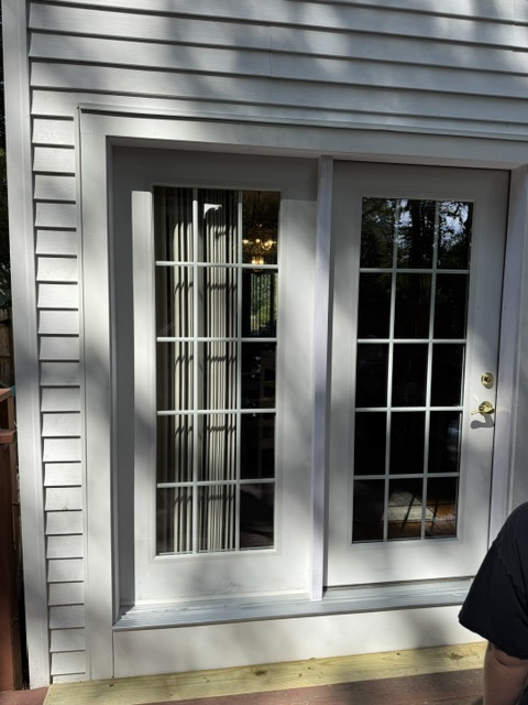 A freshly repaired front door of a New England-style home in Derry, NH, showcasing a new satin-nickel deadbolt and handle set installed perfectly straight on a smooth, freshly painted deep blue door. The crisp white trim is caulked cleanly with no gaps, and a sturdy door sweep is visible at the bottom, sealing tightly. The siding around the frame looks intact and well-maintained. Late afternoon natural light illuminates the entryway, creating soft, warm highlights on the hardware and gentle shadows on the doorstep. Photographic realism from a slightly angled close-up emphasizes precision and craftsmanship, with the surrounding porch and shrubs softly blurred to convey a sense of security, care, and attention to detail for home repair services.